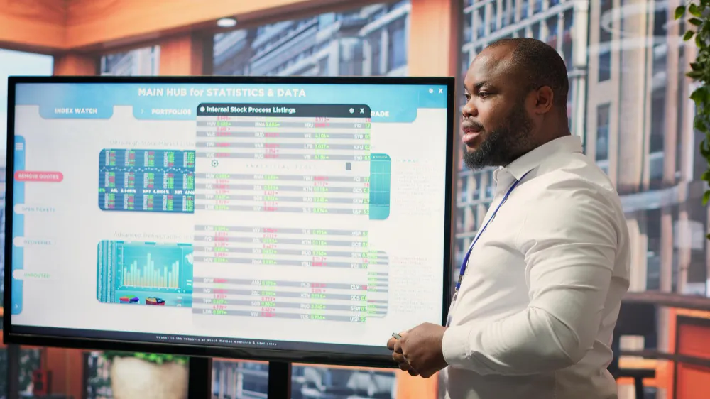 A man wearing a lanyard presents a large screen of stock figures and charts